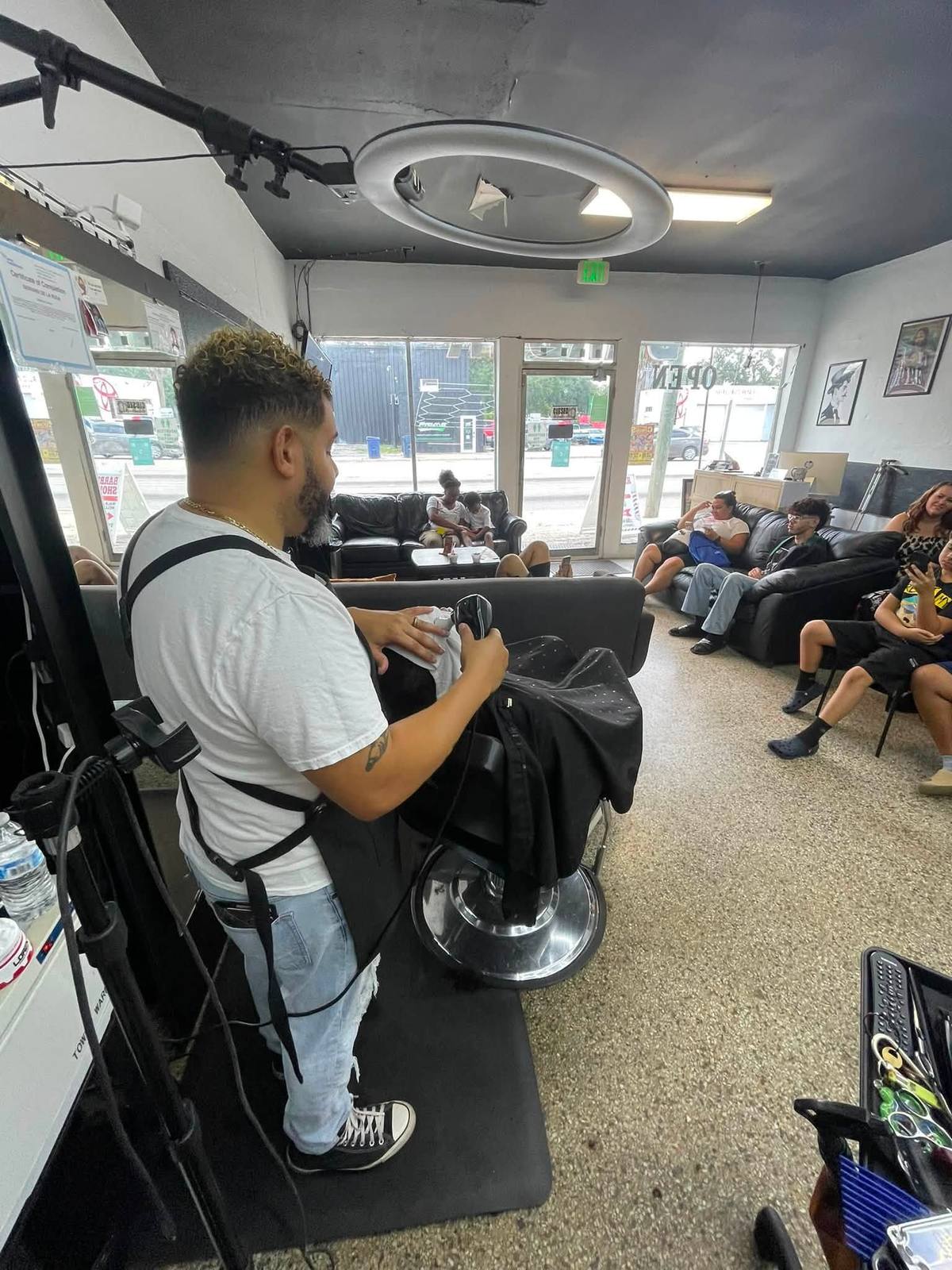 A barber at his station in a white tee and black apron, gold chain, curly blonde-tipped top over a fade. A framed Certificate of Completion on the wall behind him reads DE LA ROSA.