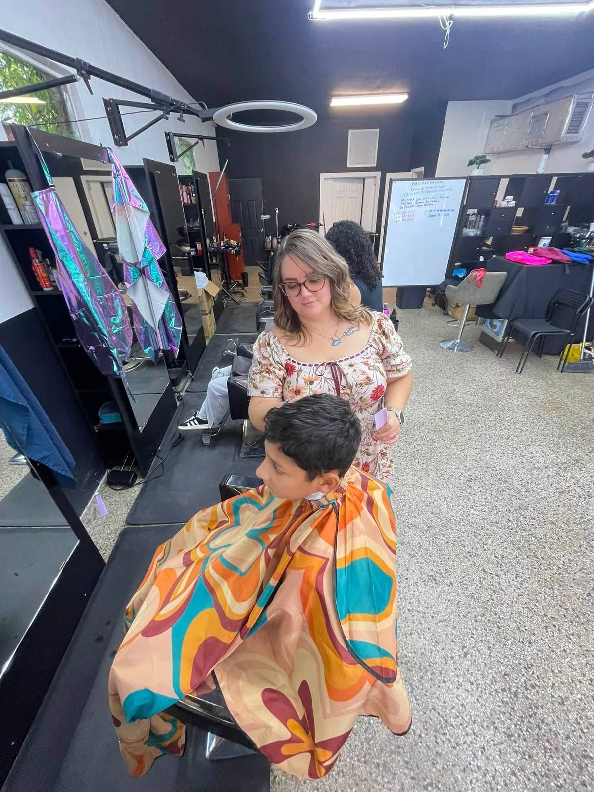 A stylist finishing a boy&rsquo;s cut, him in a retro orange-and-teal patterned cape, the shop&rsquo;s Spanish-language BIENVENIDOS service board on the back wall.