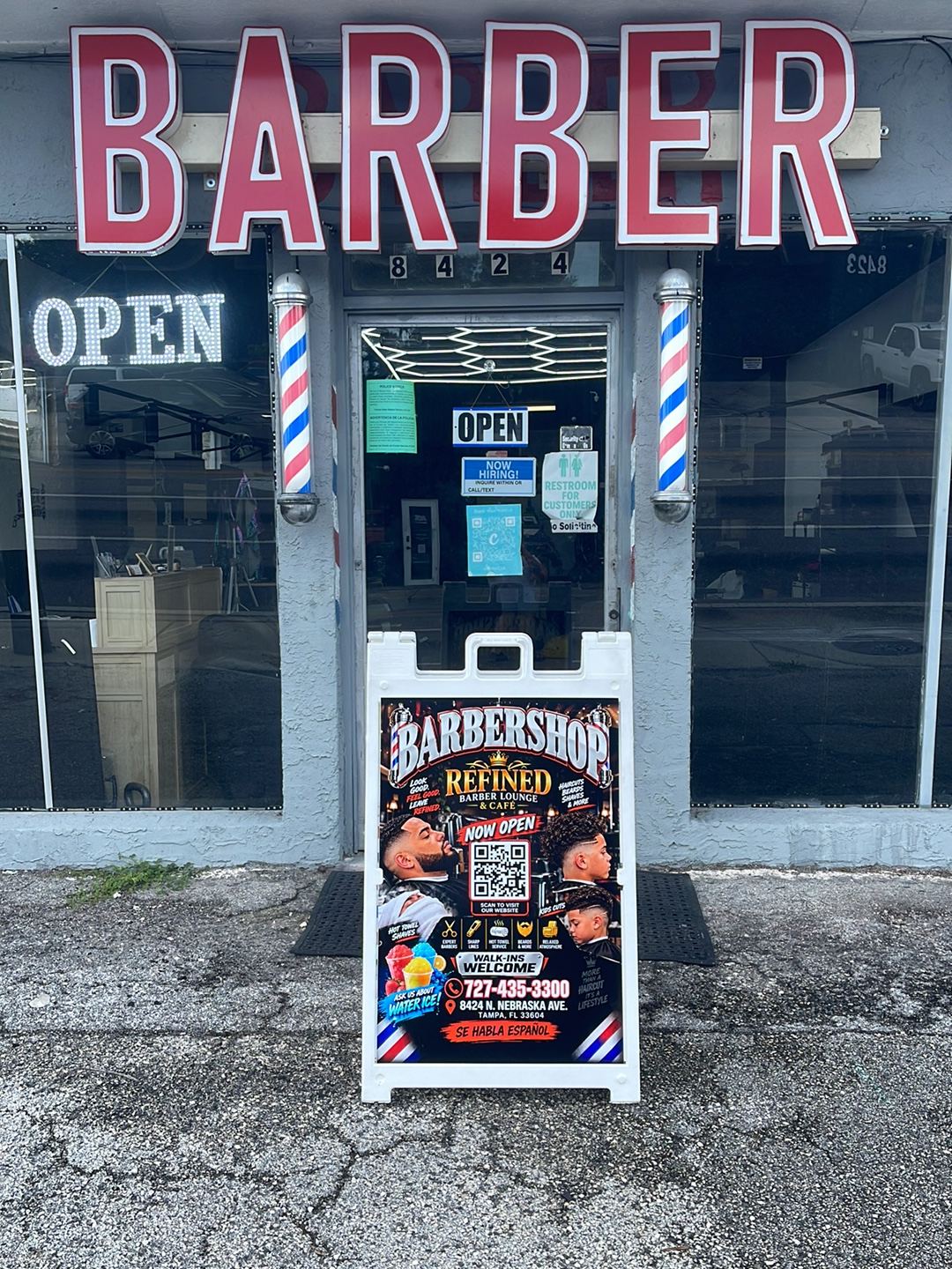 The shop from the sidewalk on N. Nebraska Ave: a red BARBER sign over the window, the 8424 door number, two barber poles either side of the door, and an A-frame sign on the pavement.